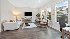 Un salon spacieux avec un canapé blanc, une table basse et un téléviseur fixé au mur. De grandes fenêtres laissent entrer la lumière naturelle et offrent une vue sur l’extérieur.