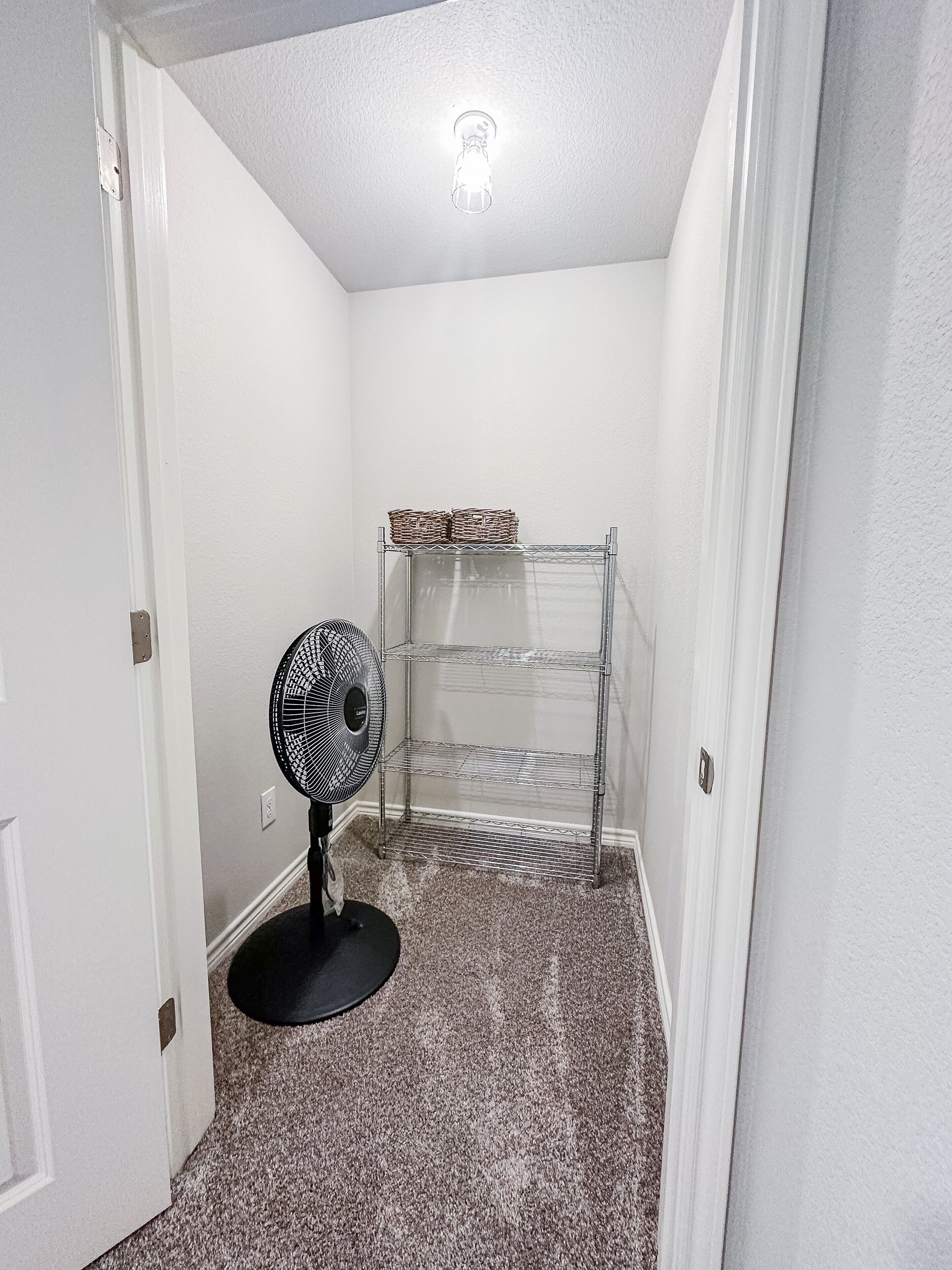 Un pasillo estrecho con un ventilador negro, una estantería metálica y una lámpara en el techo.
