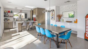 Cozinha moderna com área de refeições, destacando uma grande mesa de madeira e cadeiras azuis, uma luminária pendente e vista para o horizonte urbano pela janela.