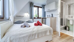 Une chambre moderne avec un plafond en pente, un lit avec des serviettes soigneusement pliées, une petite kitchenette et une salle de bain avec douche.
