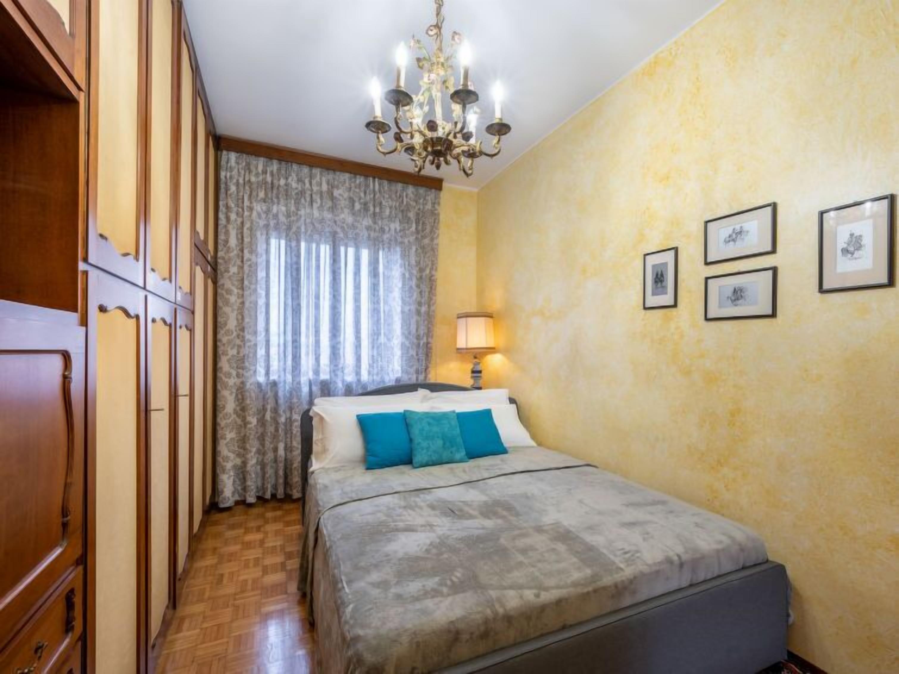 Un dormitorio con cama, candelabro, ventana con cortinas e imágenes enmarcadas en la pared.