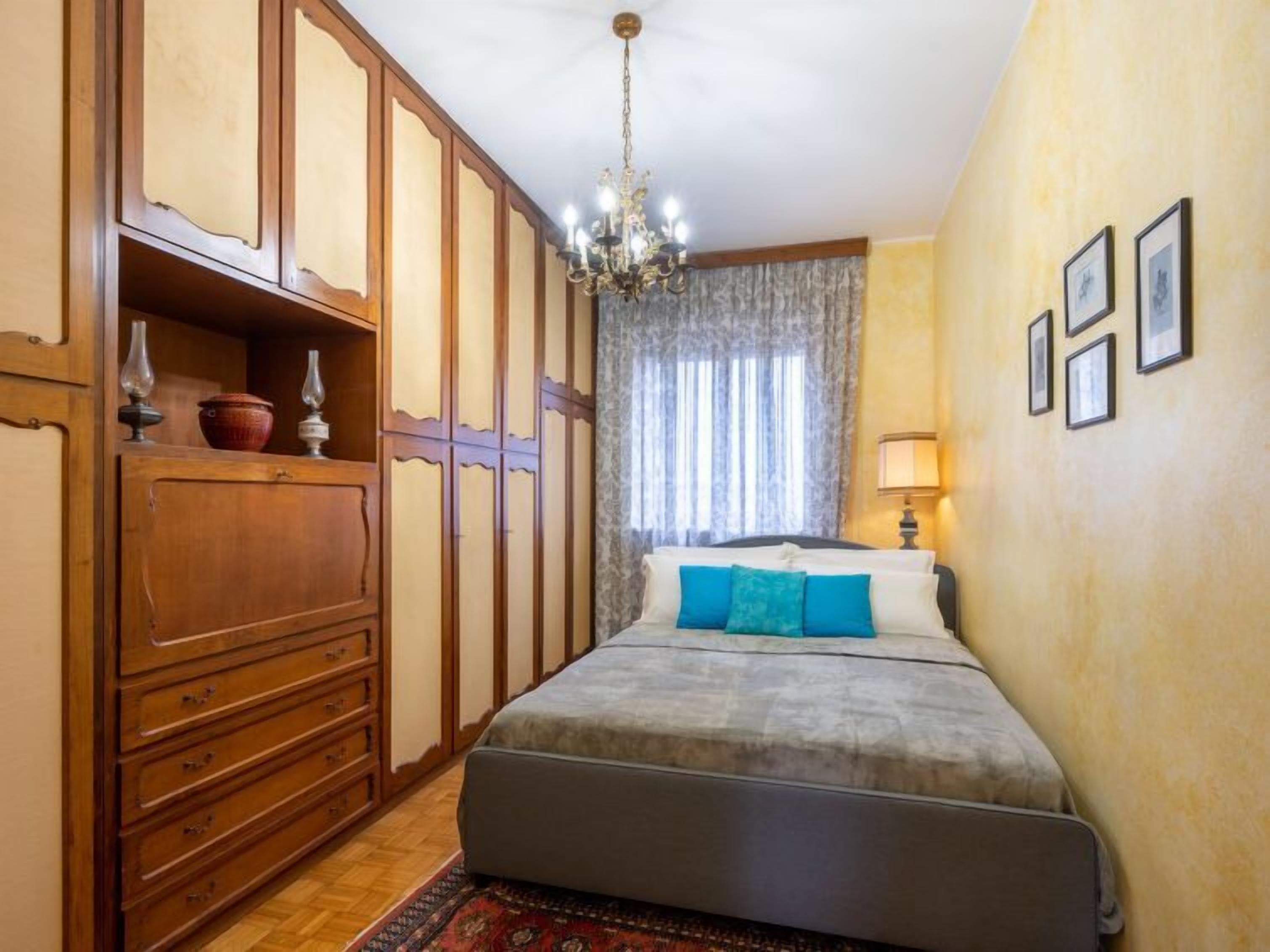 A bedroom with a bed, wooden wardrobe, chandelier, and a window with curtains.