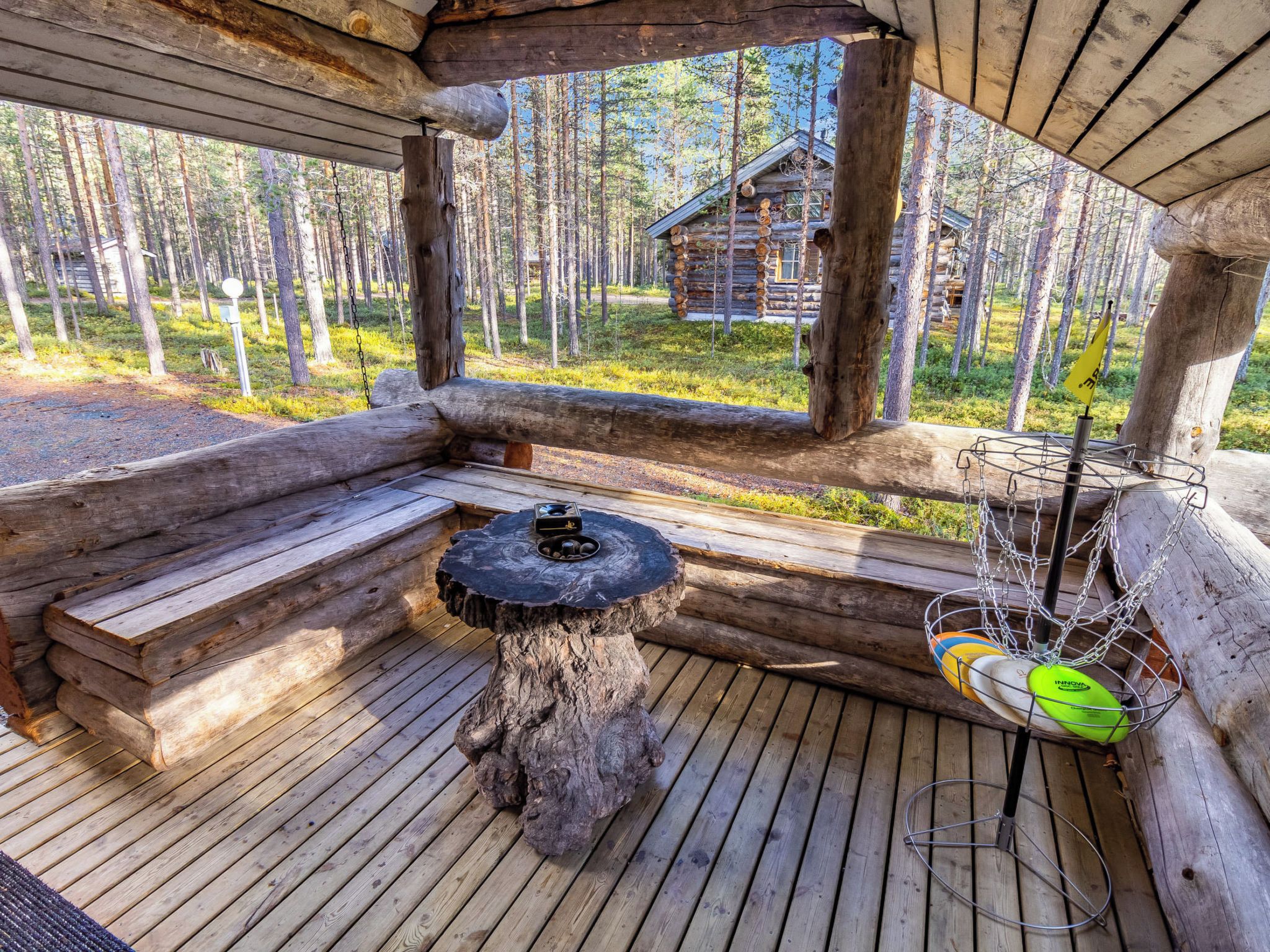 A wooden deck with a table and a hanging chair, overlooking a forest and a cabin.