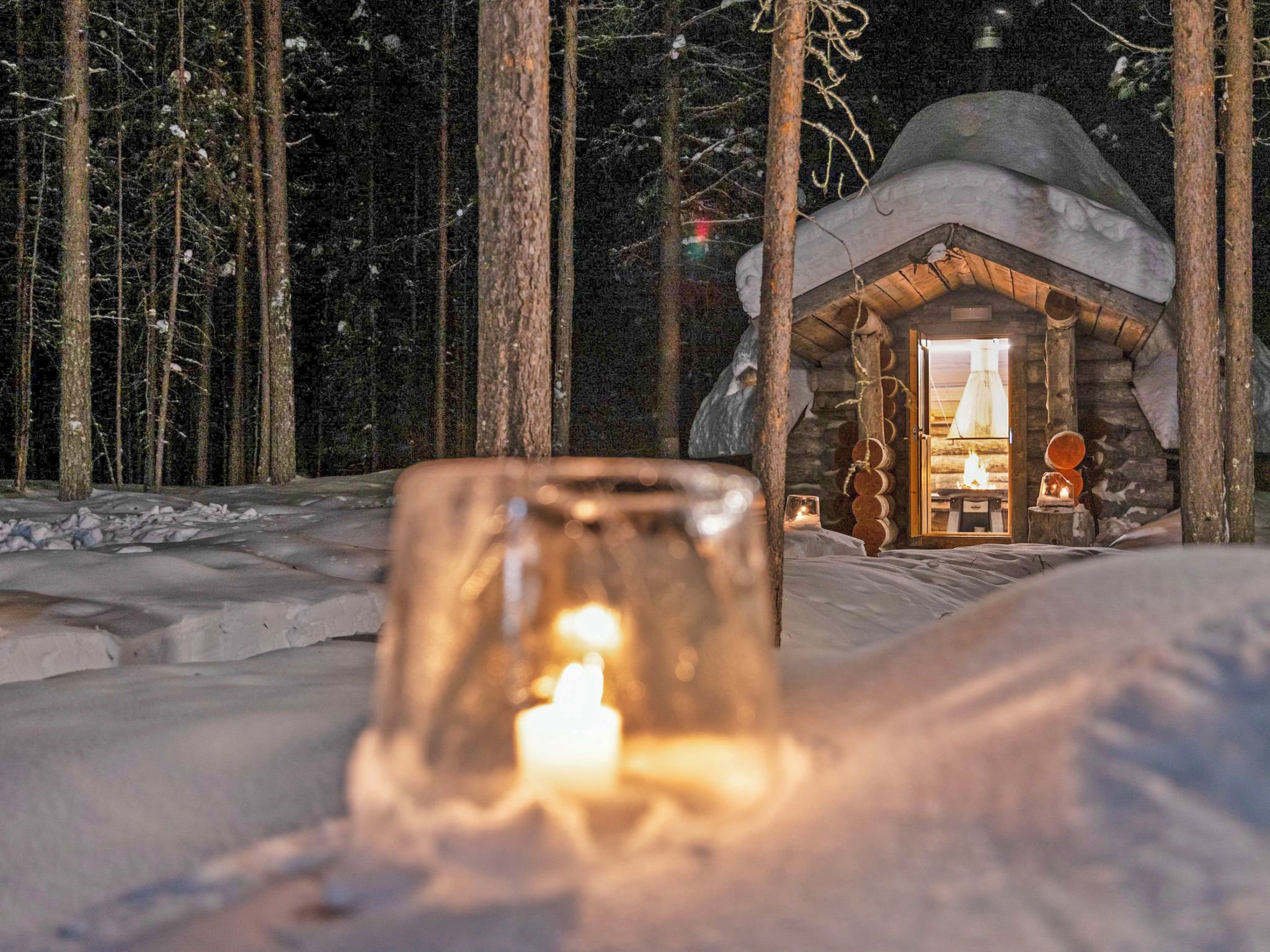 Una cabaña acogedora con una vela encendida en primer plano, rodeada de nieve en el suelo y árboles.