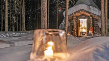 Una cabaña acogedora con una vela encendida en primer plano, rodeada de nieve en el suelo y árboles.