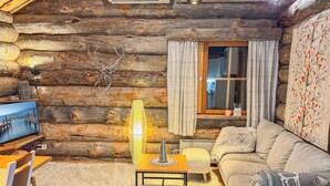 A log cabin interior with a television, a table with a lamp, a sofa, and a window with curtains.