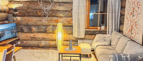 Interior de uma cabana de tronco com televisão, uma mesa com abajur, sofá e janela com cortinas.