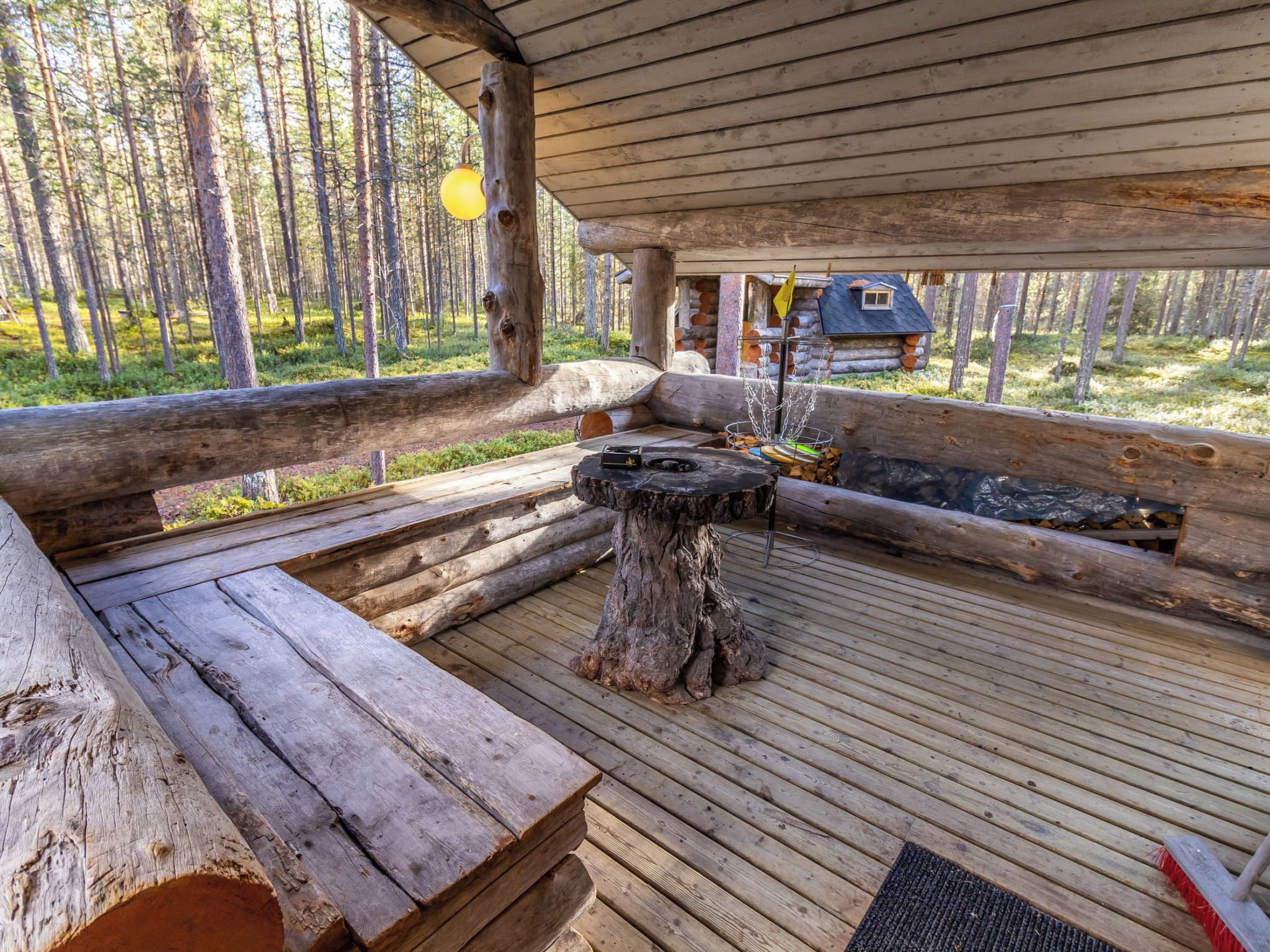 Une terrasse en bois rustique avec un foyer, des bûches et une petite cabane en arrière-plan.