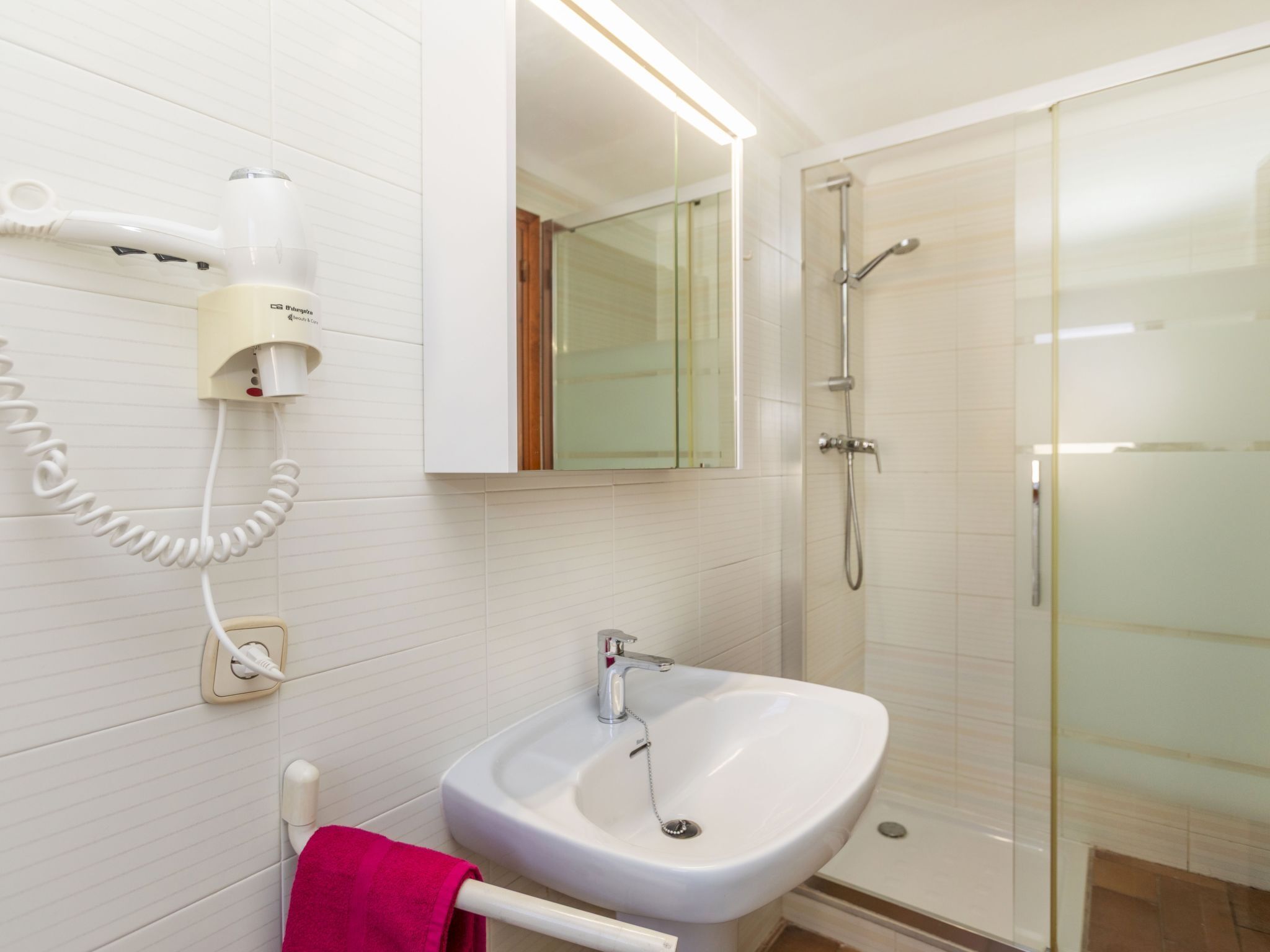 Un baño con lavamanos blanco, cabina de ducha de vidrio y un secador de pelo fijado a la pared.