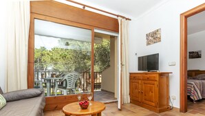 Une pièce comprenant un canapé, une table en bois avec un saladier de fruits, un téléviseur posé sur un meuble en bois, et un balcon avec une table et des chaises.