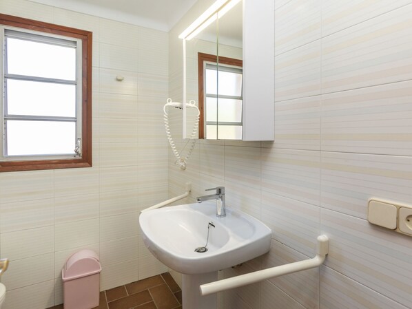 Une salle de bain avec un lavabo blanc, un miroir, une fenêtre et une poubelle rose.