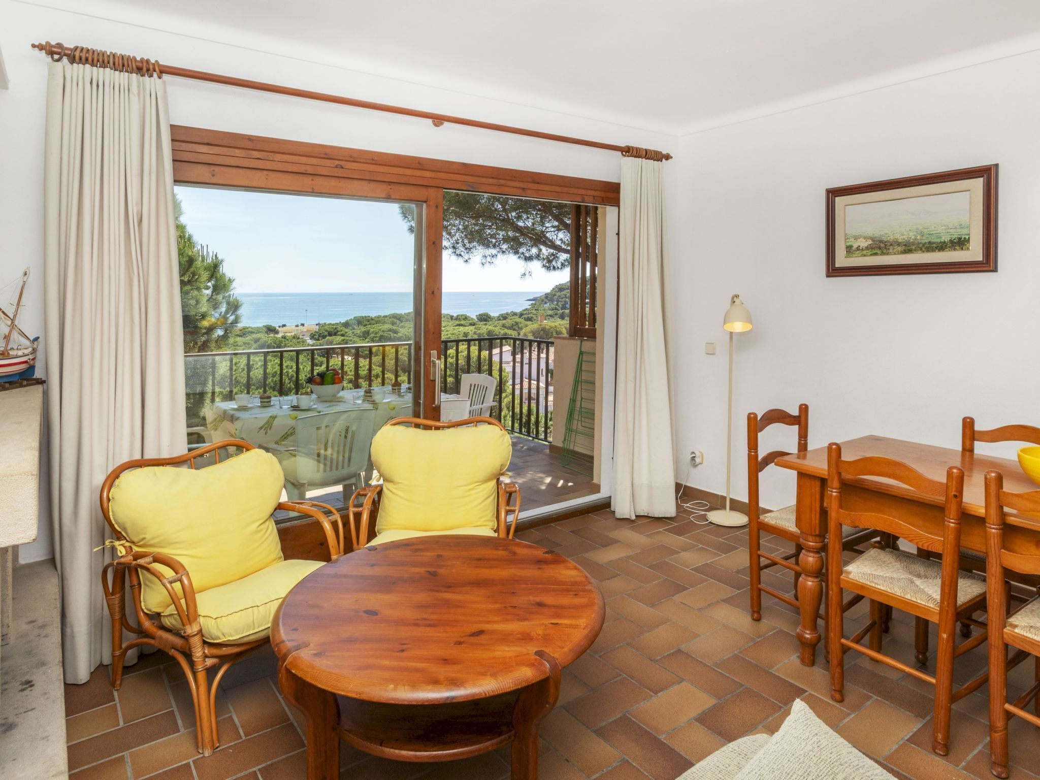 A dining area with a round wooden table and chairs, a view of the sea through a sliding glass door, and a framed picture on the wall.