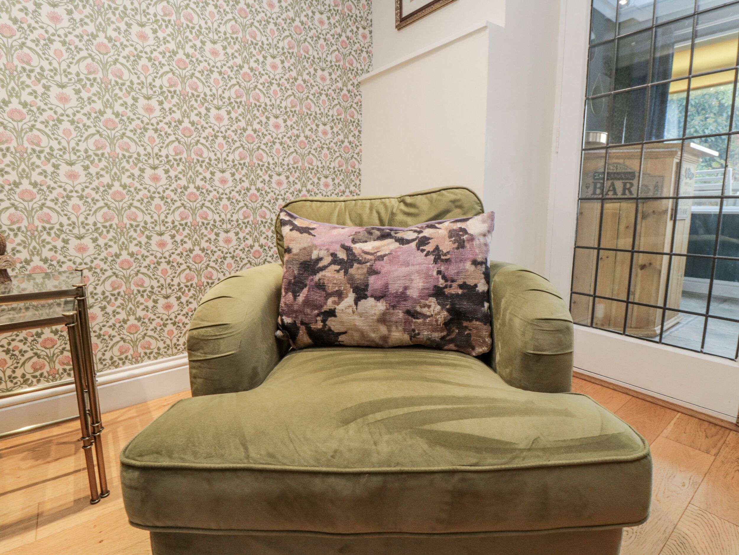 A green armchair with a floral pillow in a room with patterned wallpaper and a window with a metal grid.