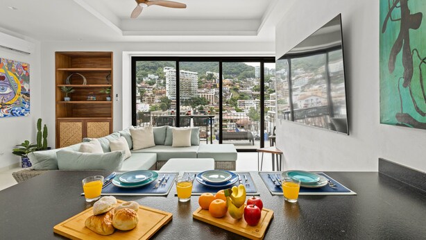 Un salon moderne avec un canapé, une table à manger où l’on prend le petit-déjeuner, une vue sur la ville et des œuvres d’art.