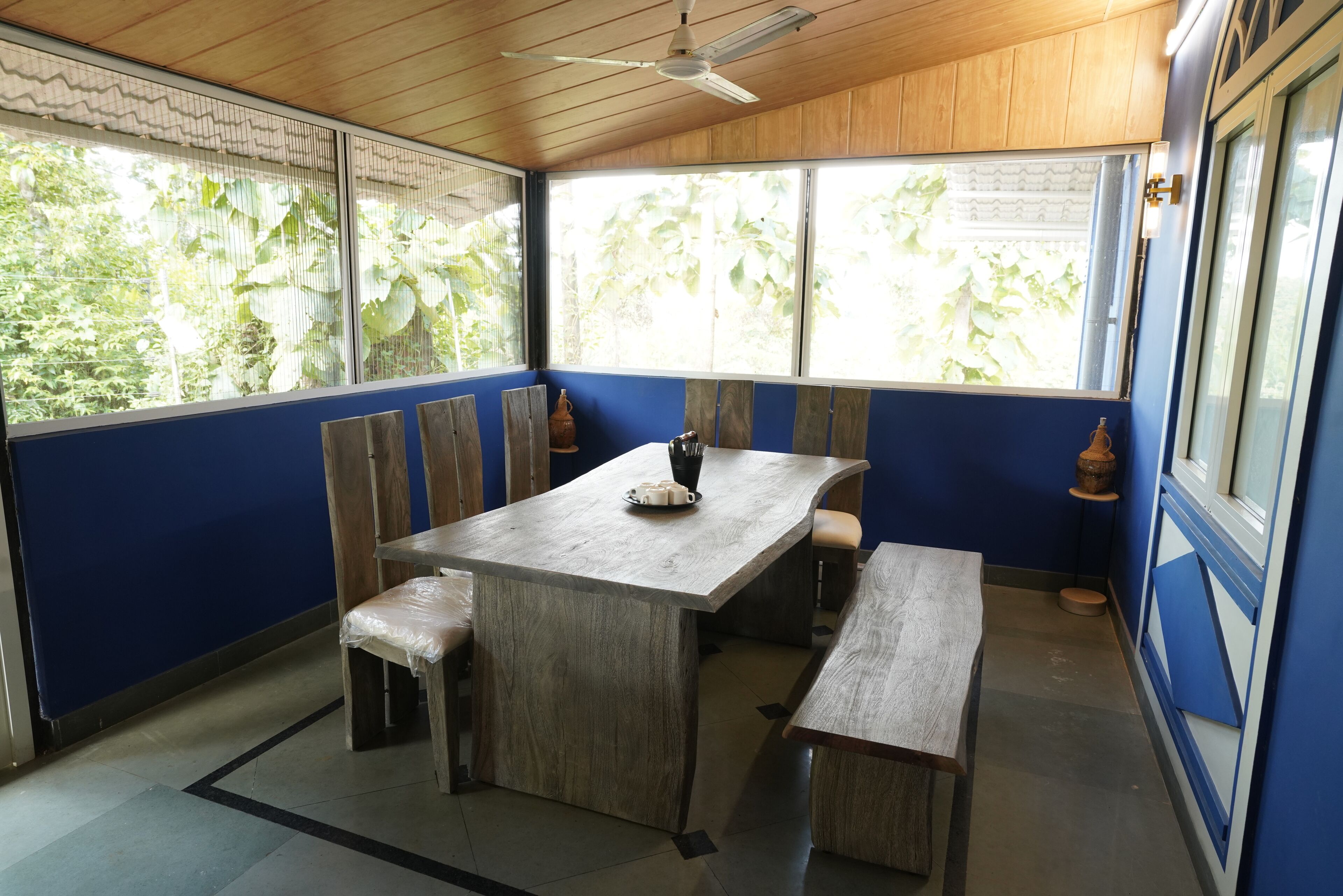A room with a wooden ceiling, a large window with a view of greenery, a table with benches, and a small rug on the floor.