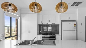 Een moderne keuken met witte kasten, een roestvrijstalen spoelbak, een gasfornuis en een witte koelkast. Boven het fornuis hangen drie rieten hanglampen.