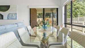 Een moderne eethoek met een glazen tafel en witte fauteuils, een blauwe vaas met bloemen en uitzicht op een omheind buitengebied.