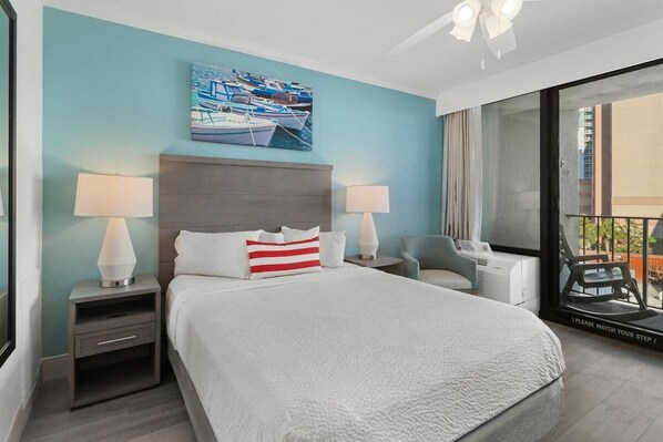 A modern hotel room with a large bed, two bedside tables with lamps, a view of the balcony, and a painting of boats on the wall.