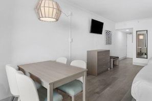 A modern hotel room with a wooden dining table, white chairs, a flat-screen TV, a wardrobe, and a mirror.