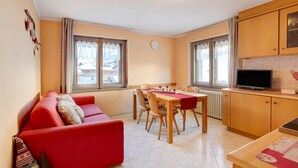 A living room with a red sofa, a dining table with chairs, a television, and a kitchen area with cabinets. - Apartment 'Lovin Winter' with Mountain View, Private Balcony and Wi-Fi (Livigno)