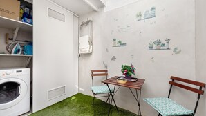 Une buanderie équipée d’un lave-linge, d’un étendoir et d’une planche à repasser. On y trouve une petite table avec un pot de fleurs et deux chaises.