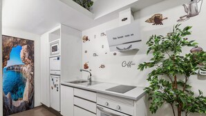 Une cuisine moderne avec des meubles blancs, un four encastré, un évier et un four à micro-ondes. Les murs sont décorés d’autocollants représentant des objets liés au café, et un grand tableau représente un paysage côtier.