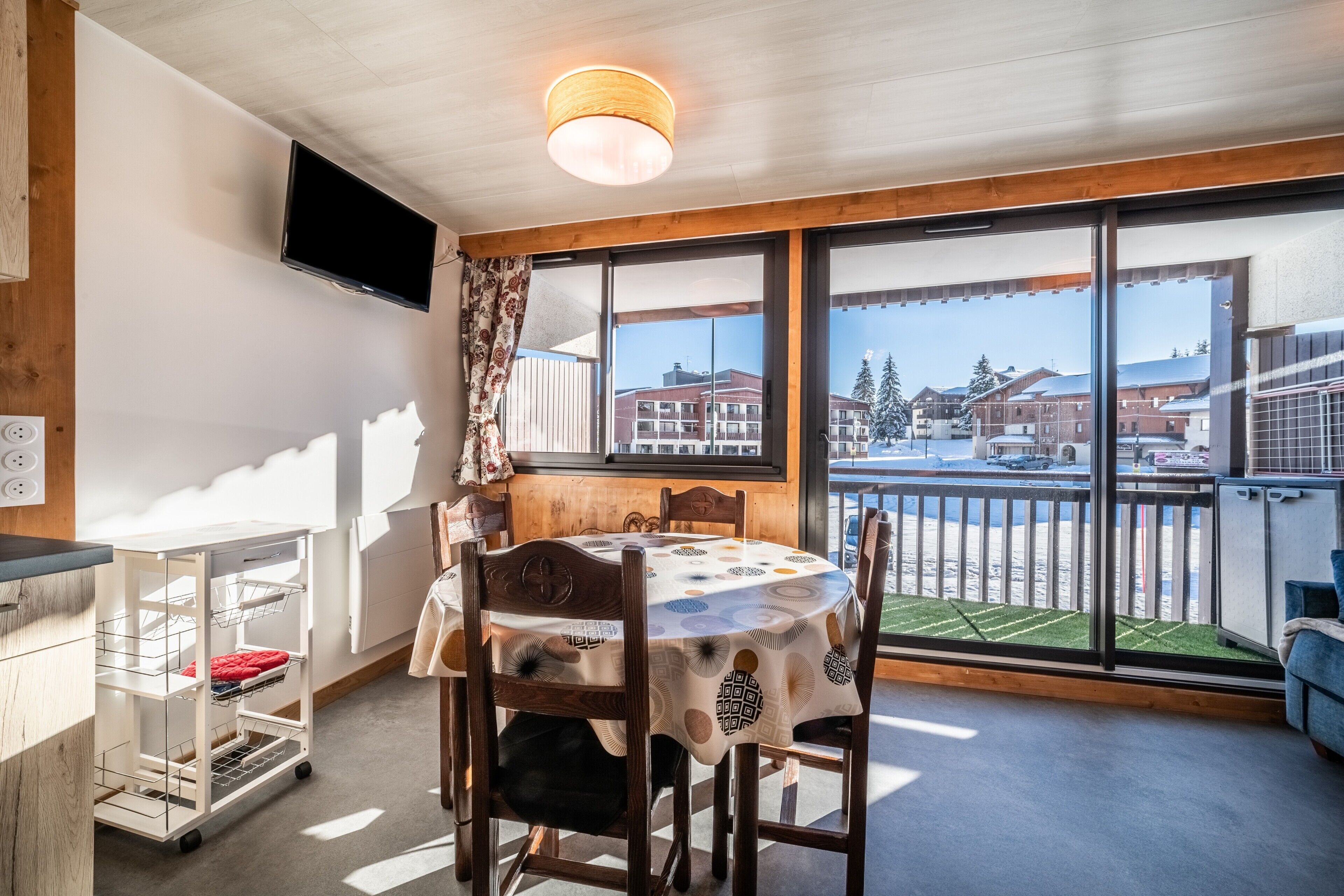 A dining area with a round table, chairs, a TV mounted on the wall, and a view of buildings through large windows.