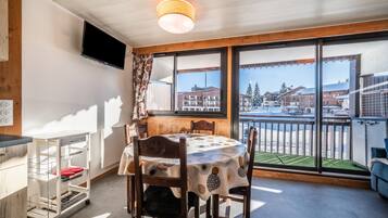 A dining area with a round table, chairs, a TV mounted on the wall, and a view of buildings through large windows.