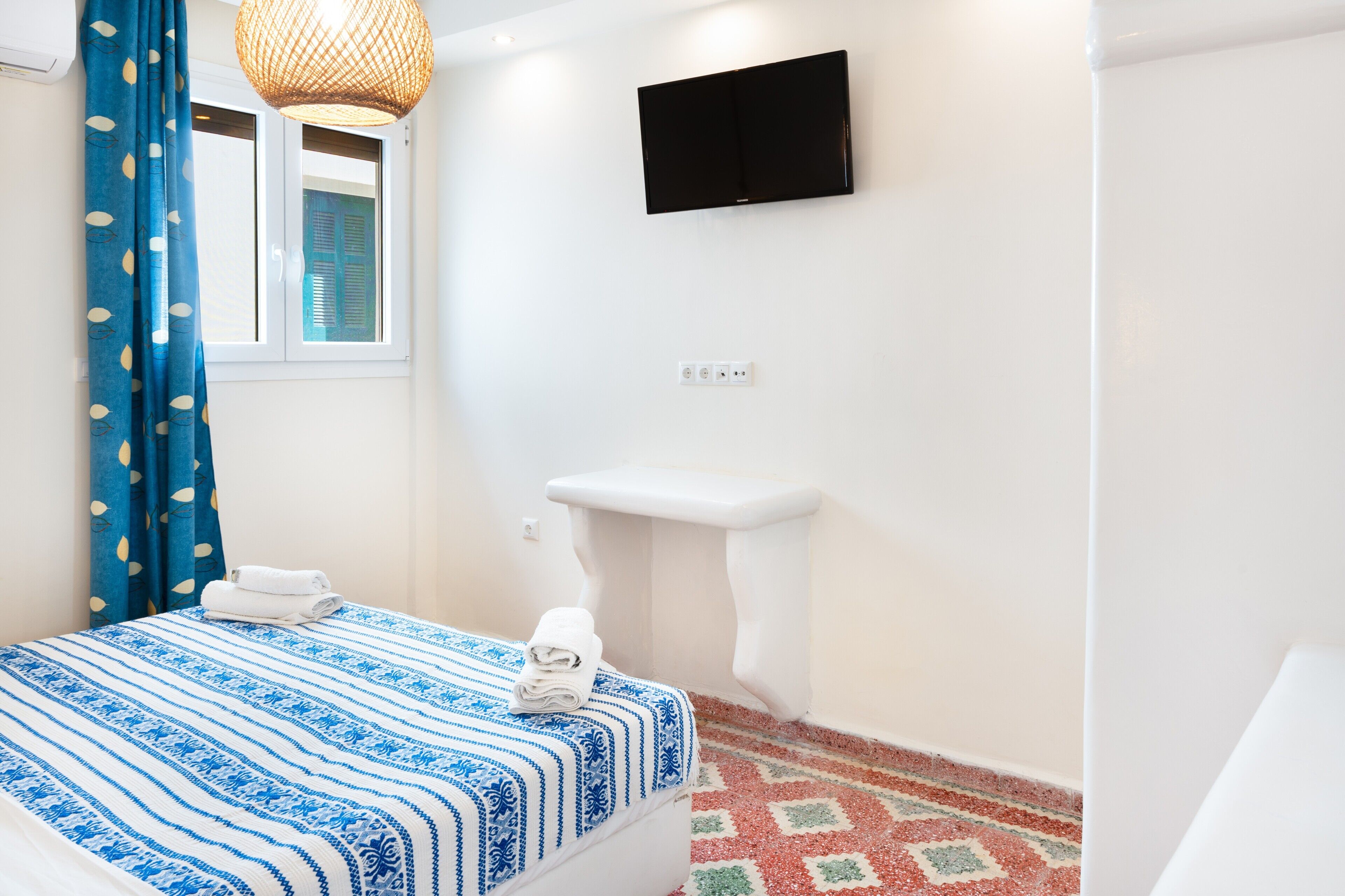 A bedroom with a bed, a window with curtains, a mounted TV, a white sink, and a patterned floor.