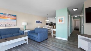 Een moderne woonkamer met een bank, fauteuil en salontafel. Er is een eethoek met een tafel en stoelen, en een keuken op de achtergrond.