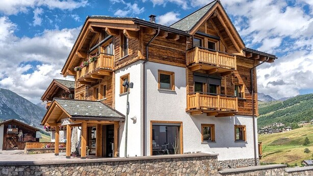 A multi-story wooden chalet with balconies, a stone foundation, and a mountainous backdrop.