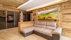 A modern kitchen with a built-in oven and refrigerator, a beige sectional sofa, and a wooden wall with a scenic mural.