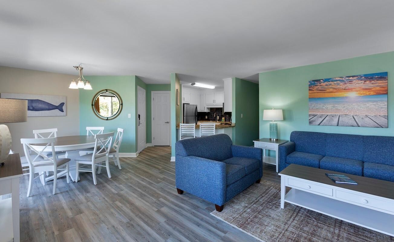 A modern living room with a blue sofa, white dining set, and a kitchen in the background.