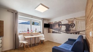 Ett modernt rum med trägolv, en blå soffa, ett skrivbord med två stolar, en tv och ett stort fönster med gardiner.