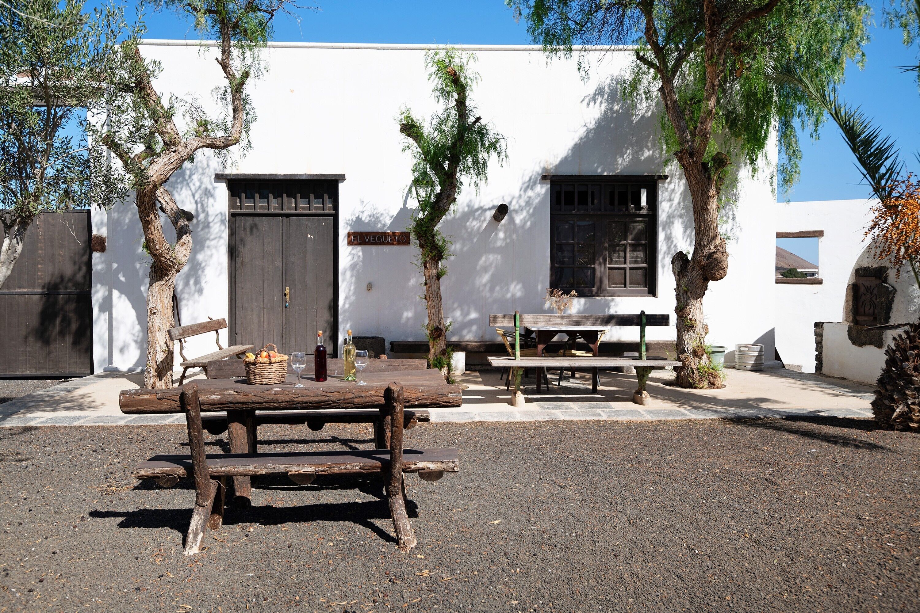 Zona salotto all'aperto dallo stile rustico, con una panca e un tavolo in legno, immersa tra alberi e un edificio bianco con una porta e finestre.