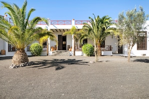 Un edificio con fachada blanca, barandal rojo en el nivel superior y un patio de grava con palmeras.