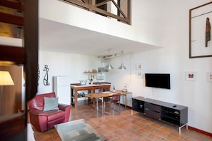 Una sala de estar con un sillón de cuero rojo, una mesa de comedor de madera, un televisor montado en la pared y una zona de cocina al fondo.