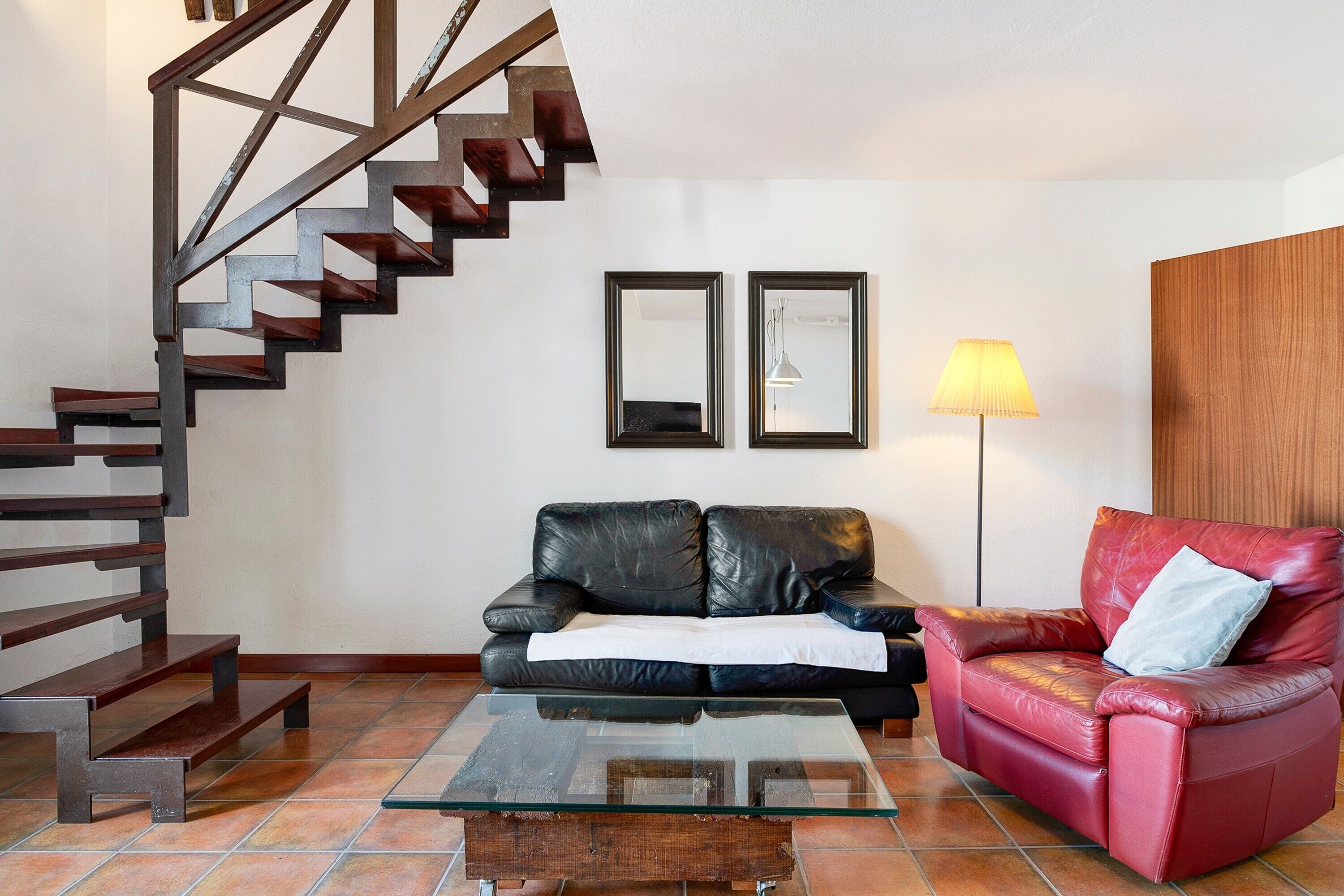 A staircase with wooden steps and metal railings, a black leather sofa, a red leather armchair, a glass coffee table, and two framed mirrors on the wall.