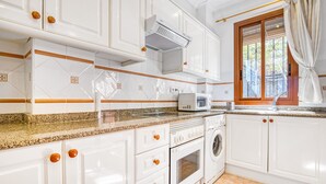 Une cuisine avec des meubles blancs, un four à micro-ondes, un lave-linge et une fenêtre avec des rideaux.