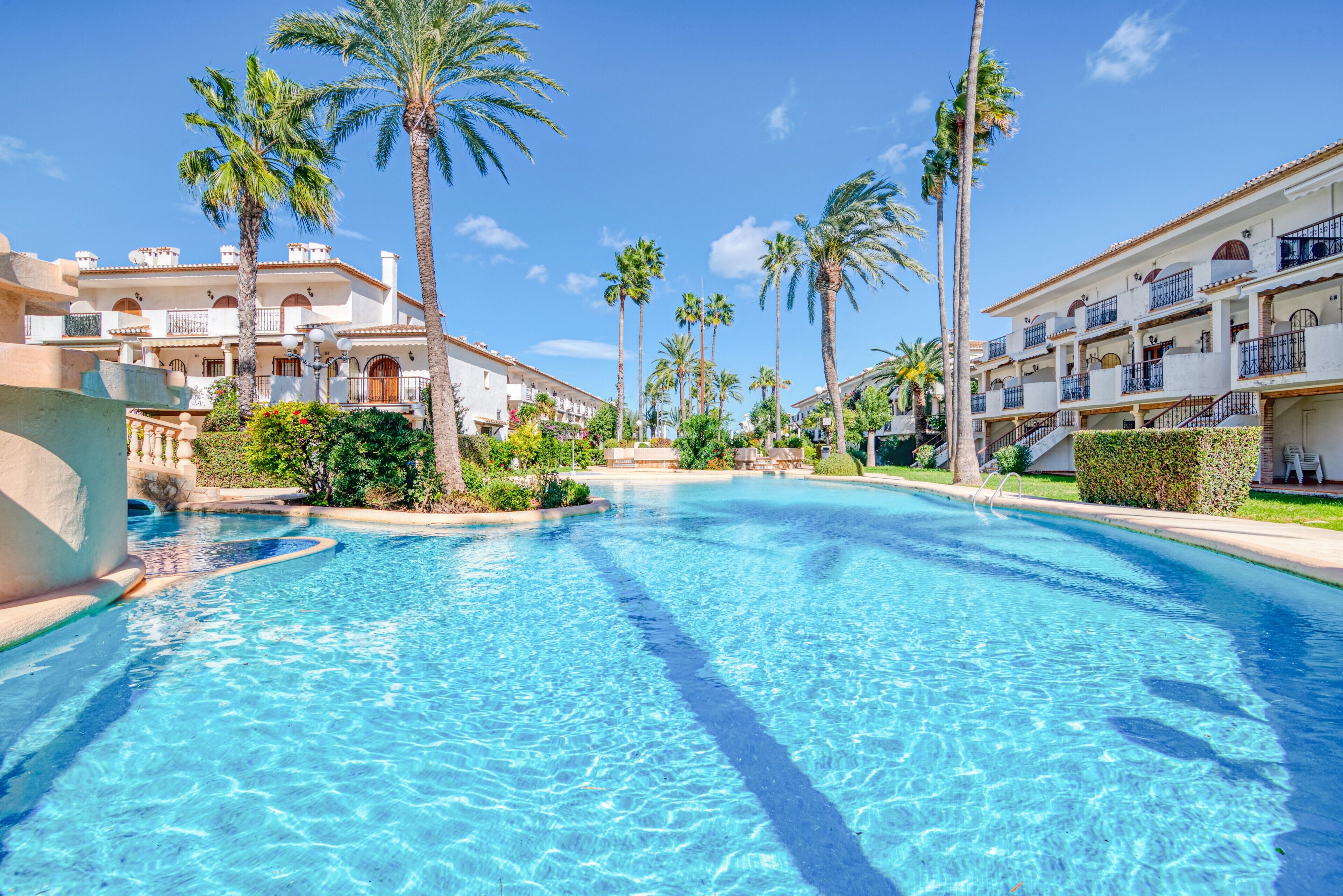 A swimming pool with clear blue water, surrounded by palm trees and multi-story buildings with balconies.