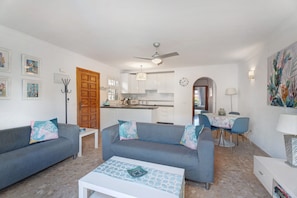 Un salon comprenant un canapé, une table basse et un coin repas avec des chaises. Au fond, on aperçoit une cuisine avec des meubles blancs et un ventilateur de plafond.
