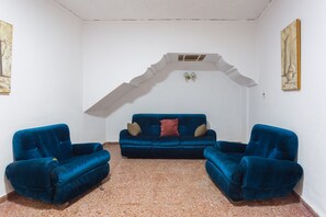 Ein Zimmer mit einem blauen Sofa, zwei gleichgestimmten Sesseln, einer weißen Decke mit einem Zierbogen und einem Gemälde an der Wand.
