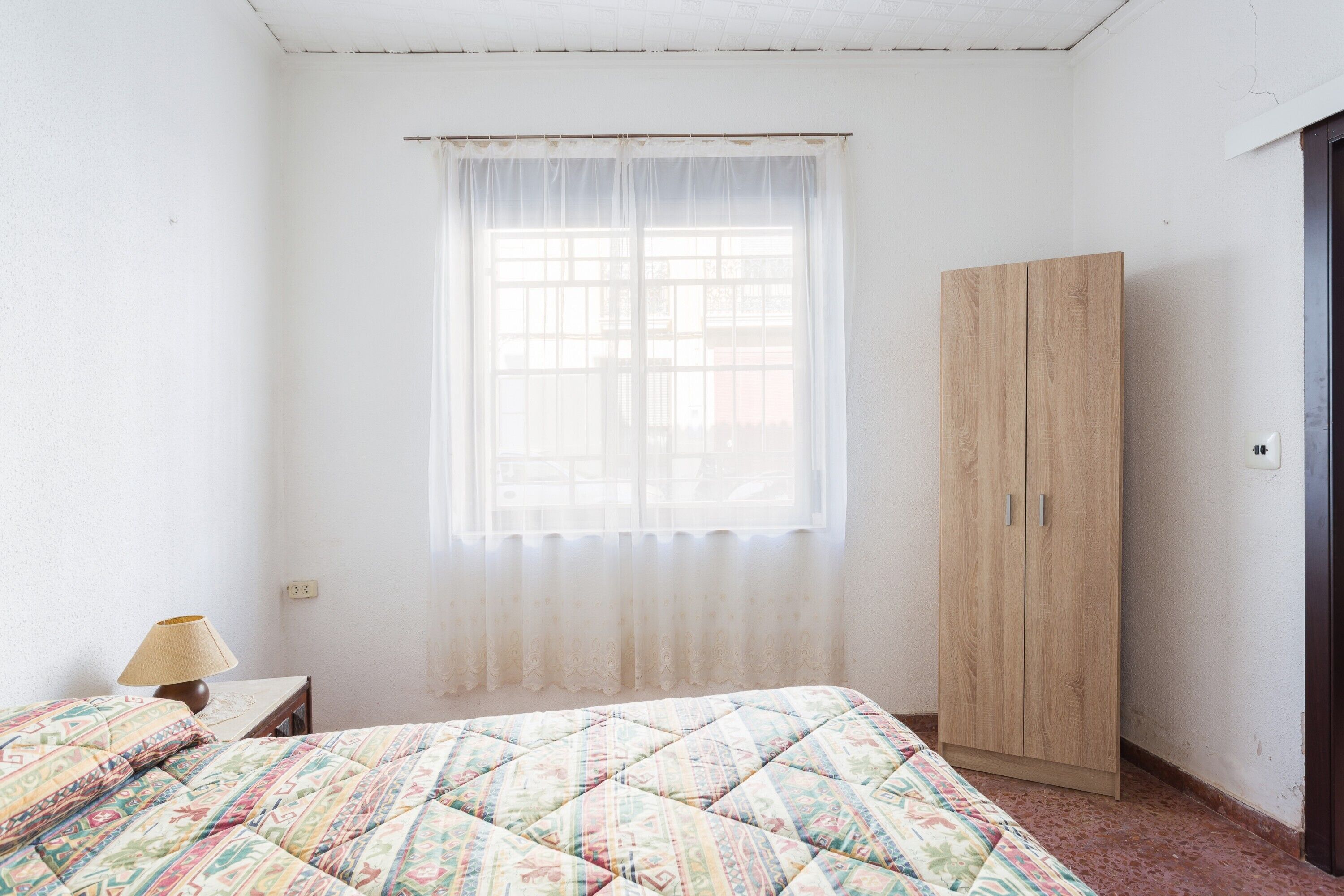 A bedroom with a bed, a window with sheer curtains, a wooden wardrobe, and a bedside table with a lamp.