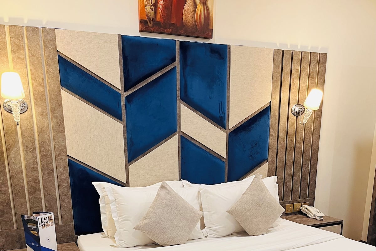 A neatly made bed with white and blue bedding, a wooden headboard with geometric patterns, and a wall-mounted artwork above.