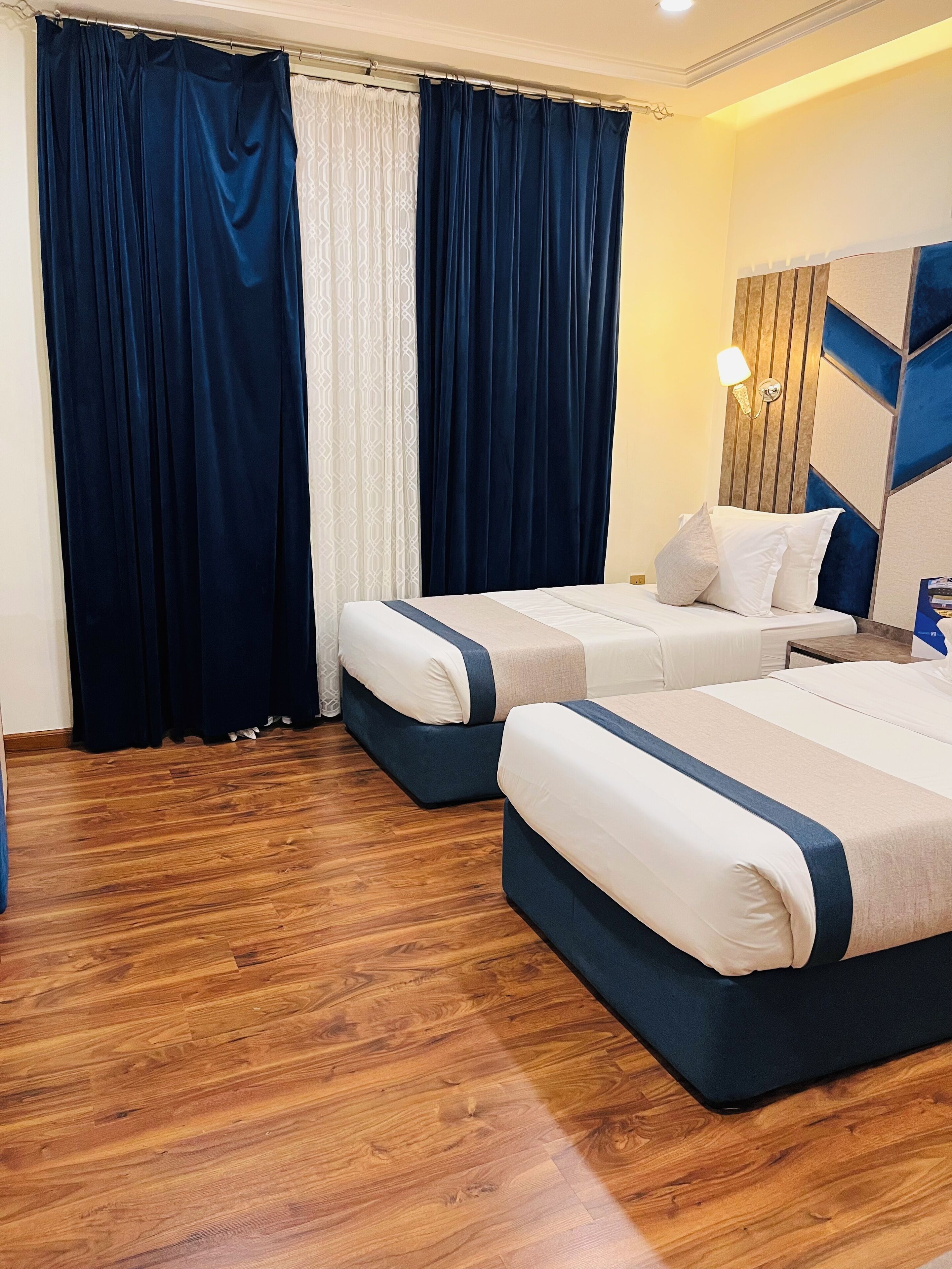 Ein Hotelzimmer mit zwei Betten, Holzboden, einem großen Fenster mit Vorhängen und einer wandmontierten Lampe.