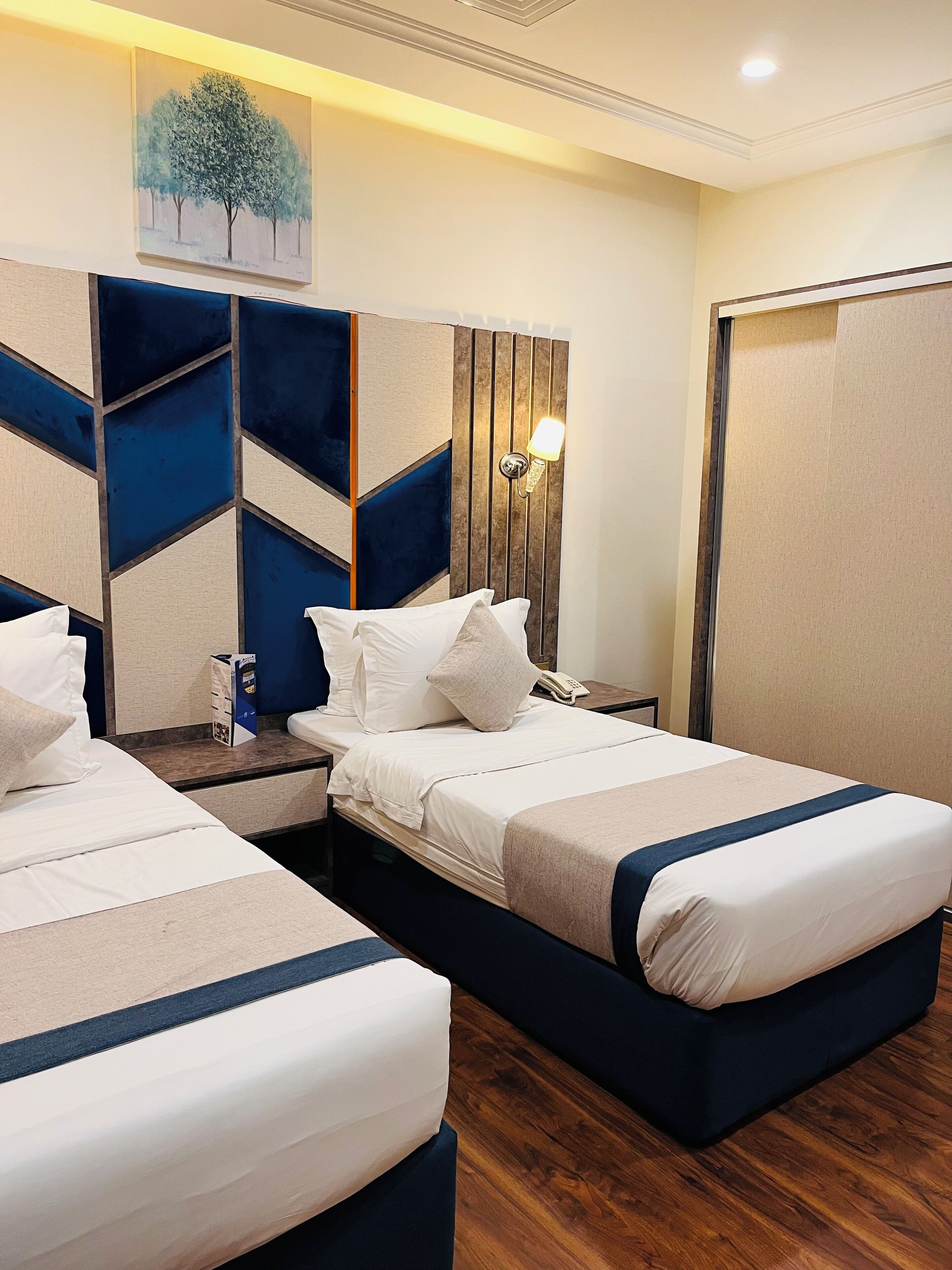A hotel room with two beds, a wooden headboard, a painting, a nightstand, and a wall-mounted lamp.