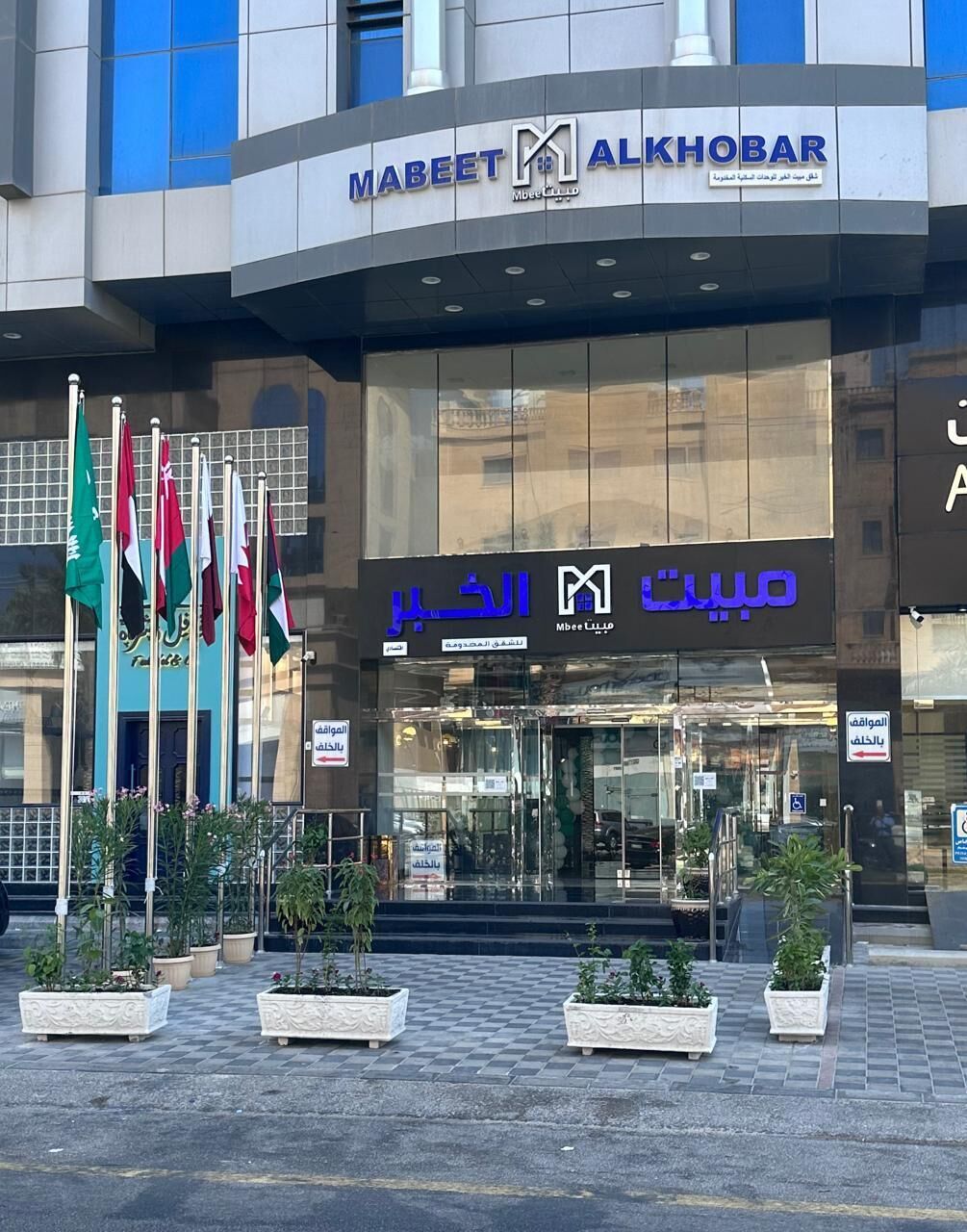 Un edificio moderno con un cartel que dice 'Mabeeet Alkhoobar' y 'Mbee' en árabe. Hay varias banderas al frente, y macetas con plantas decoran la entrada.