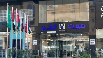 Un edificio moderno con un cartel que dice 'Mabeeet Alkhoobar' y 'Mbee' en árabe. Hay varias banderas al frente, y macetas con plantas decoran la entrada.