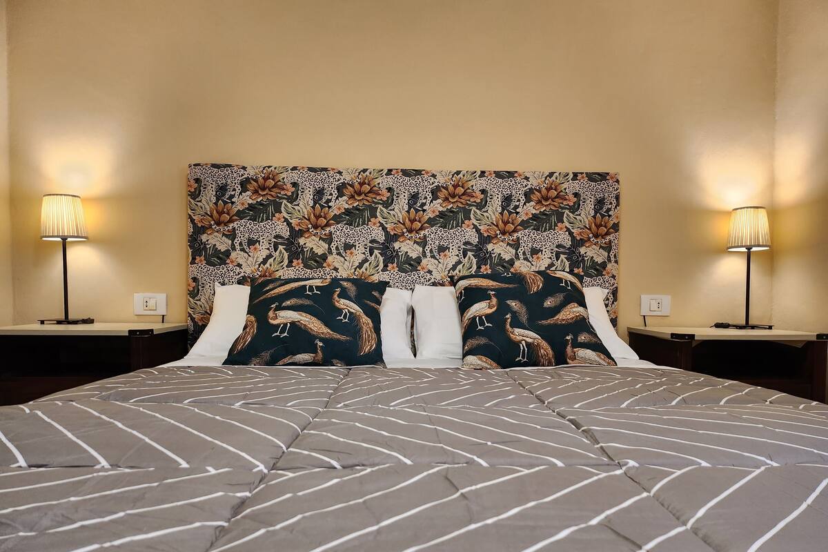Een bed met een bedsprei met patroon, twee decoratieve kussens en een hoofdbord met bloemendessin. Aan weerszijden van het bed staan twee bedlampen.