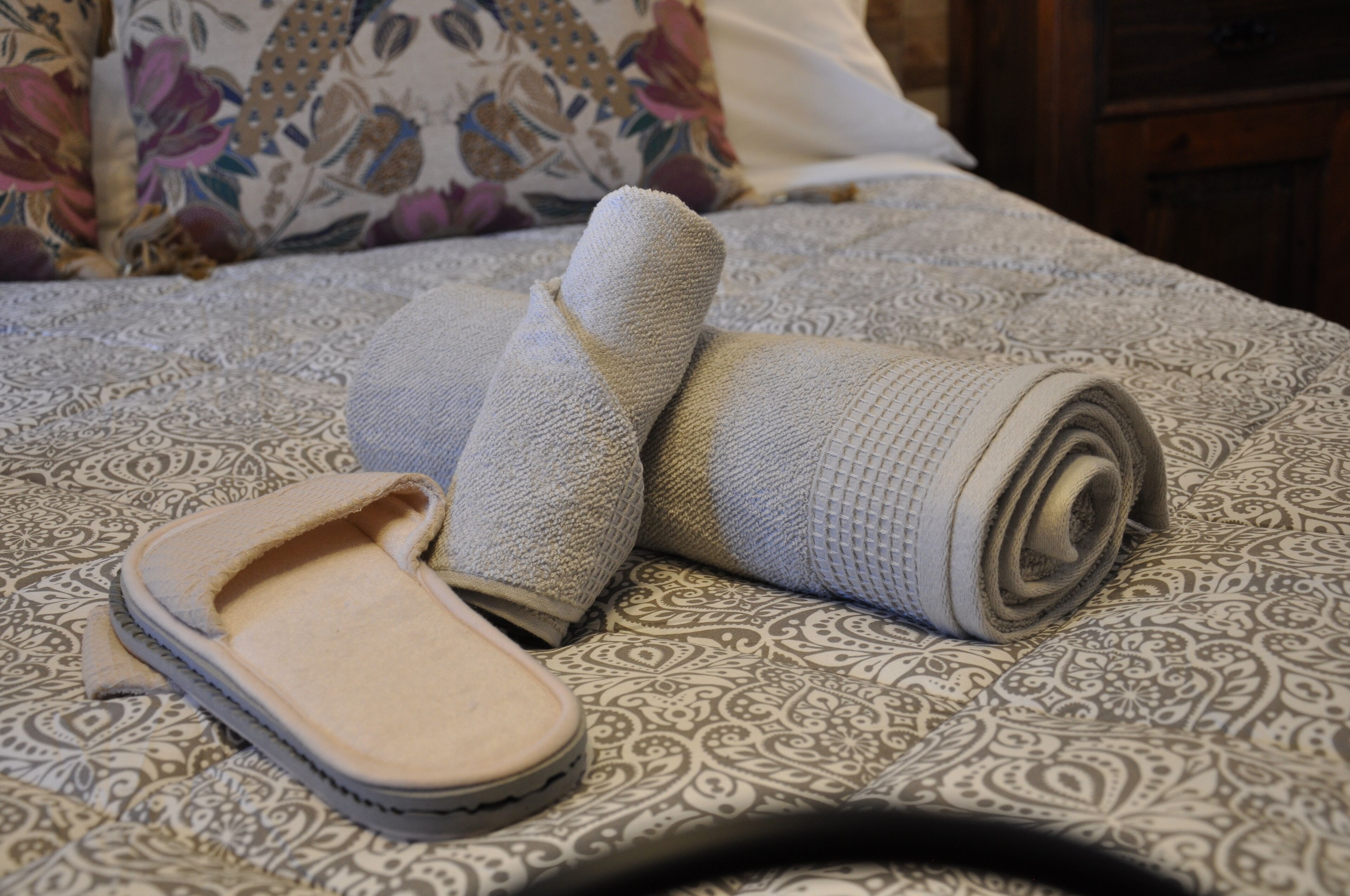 Ein Bett mit einem gemusterten Überwurf, ein Paar Hausschuhe und ein aufgerolltes Handtuch.